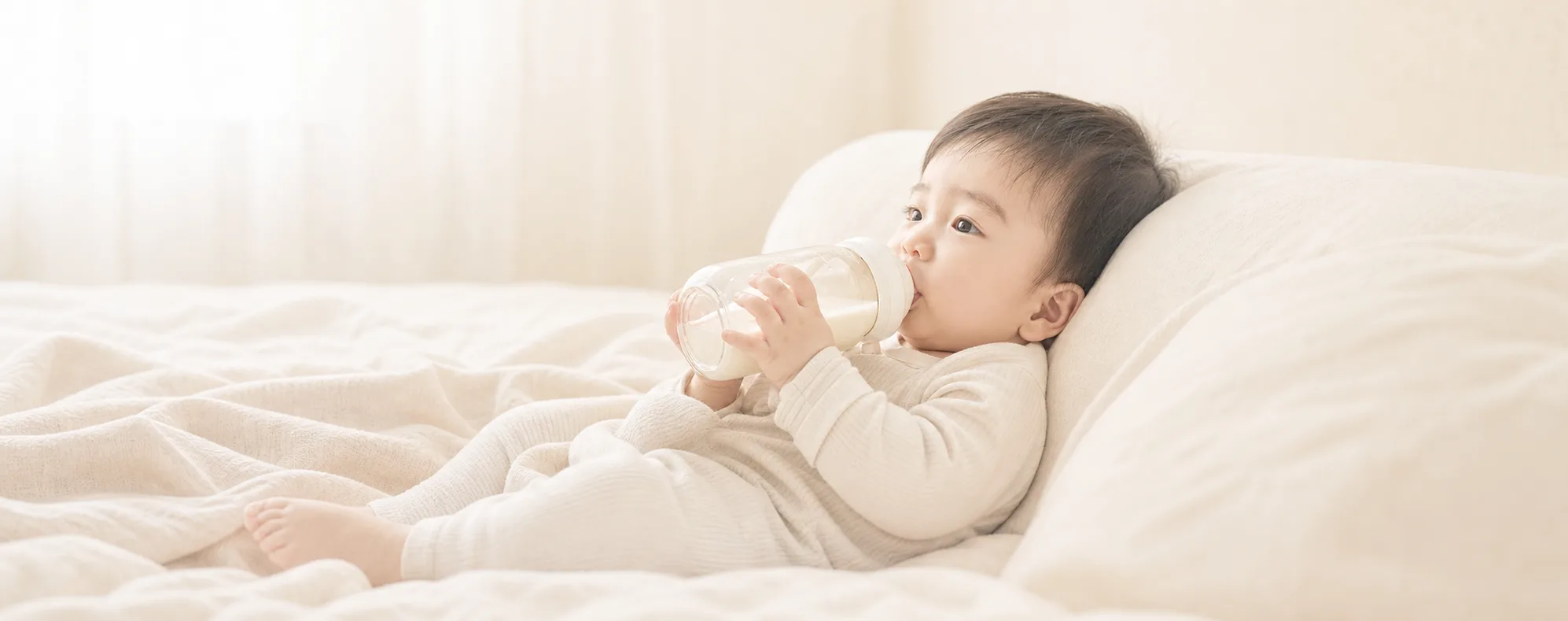 밝은 아이보리 침구에 기대어 젖병을 먹는 아기